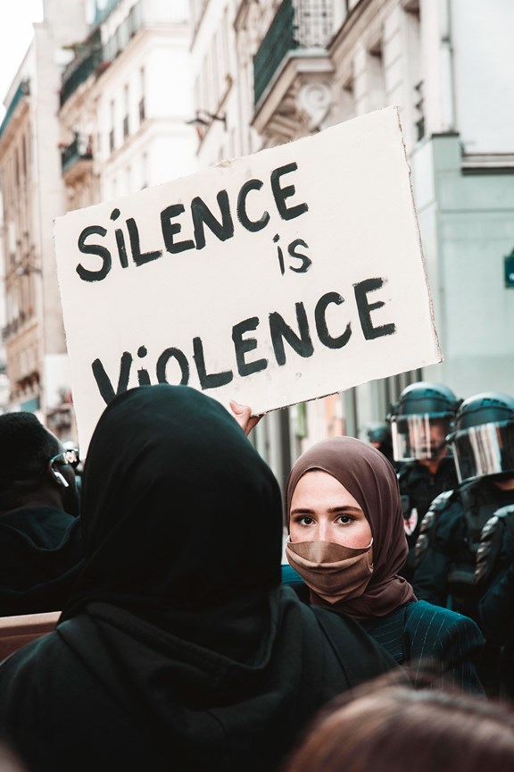 Frau mit Kopftuch und Mund-Nasen-Schutz steht in der Menge und hält Schild in die Luft mit dem Text: "Silence is Violence"